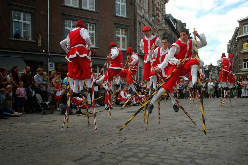 Stilt walkers in red and white costumes, with balancing poles, performing on a cobbled street